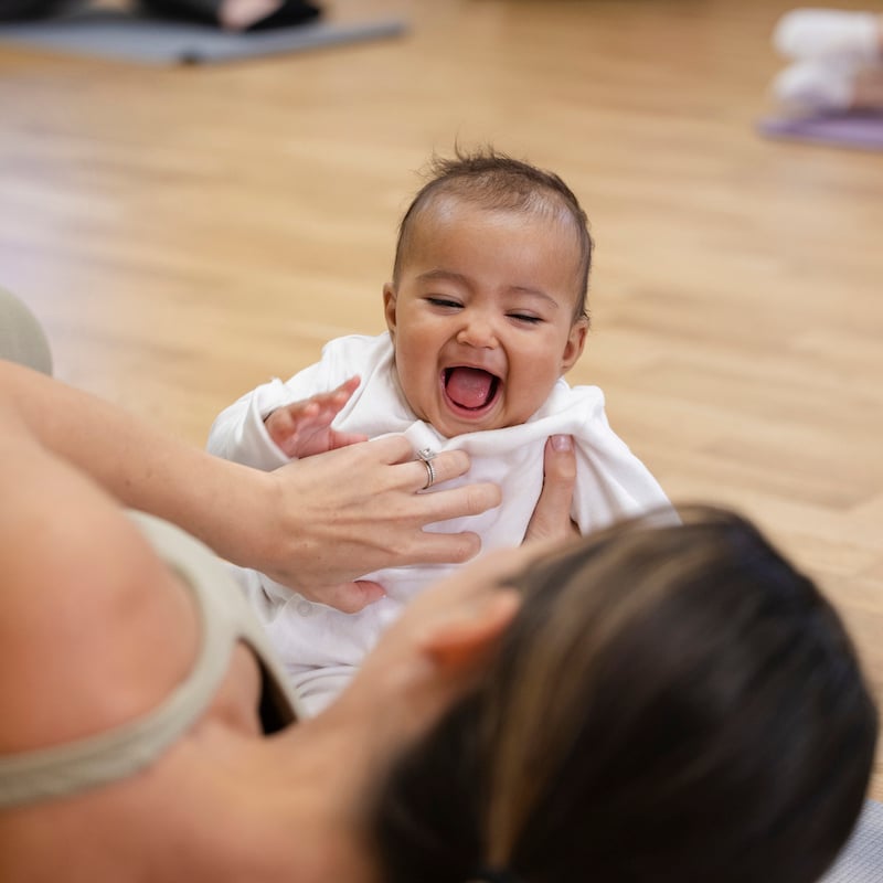 A shot of a mother in a yoga class, she is lying on her side while tickling her young baby girl on an exercise mat.